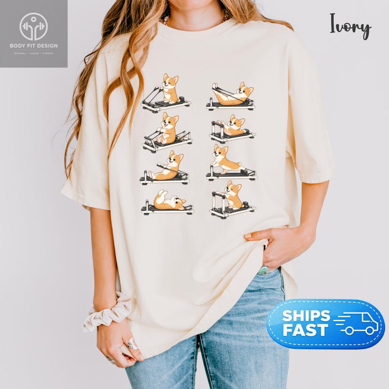 Op de afbeelding: Ivoor kleurig T-shirt met een ontwerp van cartoon corgi's die oefenen op Pilates apparatuur. De corgi's worden in verschillende poses getoond. De tekst "Body Fit Design" staat linksboven. Het shirt heeft een relaxte pasvorm.
