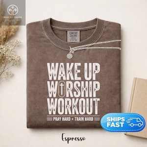 Może przedstawiać: Brązowy t-shirt z okrągłym dekoltem i białym, postarzanym napisem WAKE UP WORSHIP WORKOUT oraz PRAY HARD TRAIN HARD. Koszulka posiada metkę marki Comfort Colors przy wewnętrznym dekolcie oraz mały okrągły wisiorek spoczywający na kołnierzyku.