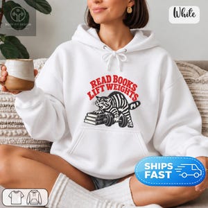 May include: A white hooded sweatshirt featuring a graphic print of a black and white striped cat lifting a dumbbell while resting its other paw on a stack of books. The red text above the cat reads READ BOOKS LIFT WEIGHTS in a bold font.