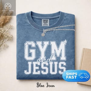 以下が含まれることがあります： GYM and JESUSという文字が太字の大学風フォントでプリントされた、ブルーのクルーネックTシャツ。かすれた白のグラフィックが特徴で、リブ編みのネックライン、内側のComfort Colorsブランドラベル、シルバーのネックレスが添えられています。