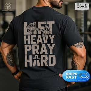 Może przedstawiać: Czarny t-shirt z krótkim rękawem z grafiką na plecach przedstawiającą napis LIFT HEAVY PRAY HARD w postarzanej beżowej czcionce. Projekt zawiera ilustrację sztangi nad tekstem oraz krzyż zastępujący literę A w słowie HARD.