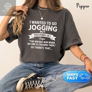 May include: A charcoal gray short-sleeve t-shirt featuring white text that reads I WANTED TO GO JOGGING BUT PROVERBS 28:1 SAYS THE WICKED RUN WHEN NO ONE IS CHASING THEM. SO THERE'S THAT. The shirt has a crew neckline and a relaxed, loose-fitting cut.