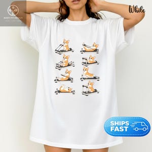 Op de afbeelding: Wit T-shirt met een ontwerp van corgi honden die pilates apparatuur gebruiken. De illustraties staan in een reeks. Het shirt heeft een ronde hals en korte mouwen. De tekst "Body Fit Design" en een logo zijn ook zichtbaar.