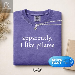 May include: A violet-colored t-shirt with the phrase "apparently, I like pilates" in white lettering. A silver necklace with a sunburst pendant is draped over the shirt. The shirt is folded neatly, and the word "Violet" is at the bottom.