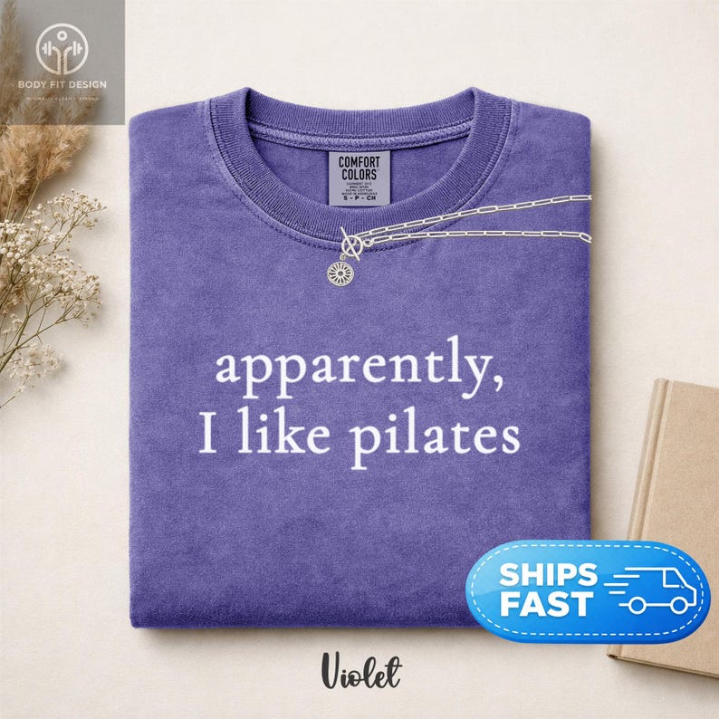 May include: A violet-colored t-shirt with the phrase "apparently, I like pilates" in white lettering. A silver necklace with a sunburst pendant is draped over the shirt. The shirt is folded neatly, and the word "Violet" is at the bottom.