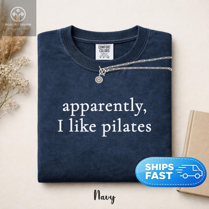 May include: Navy blue t-shirt with the text "apparently, I like pilates" in white. The shirt has a crew neck and a silver necklace with a sunburst pendant. The brand name "Comfort Colors" is visible on the tag.