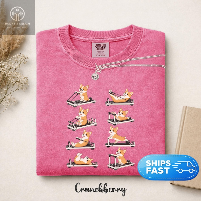 Op de afbeelding: Roze T-shirt met ronde hals en een herhaalde afbeelding van corgi-honden die oefeningen doen op pilates-reformers. De tekst "Crunchberry" staat onderaan het shirt. Een zilveren ketting met een zonnehanger ligt op het shirt.
