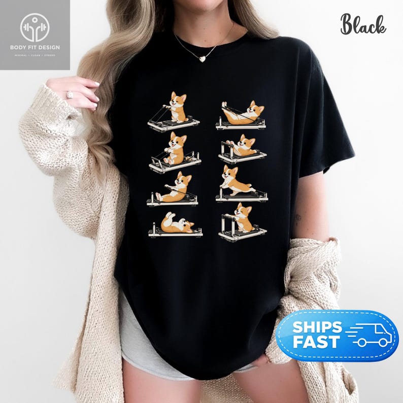 Op de afbeelding: Zwart T-shirt met een ontwerp van cartoon corgi honden die Pilates reformers gebruiken. De corgi's worden getoond in verschillende oefenposities, met oranje en witte vacht. Het shirt heeft het woord "Black" rechtsboven. Het shirt heeft ook de tekst "BODY FIT DESIGN" linksboven.