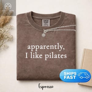 May include: Espresso-colored t-shirt with the text "apparently, I like pilates" in white. The shirt has a crew neck and a silver necklace. A blue and white "Ships Fast" sticker is on the bottom right.