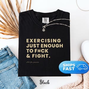May include: Black t-shirt with the text "EXERCISING JUST ENOUGH TO F≈CK & FIGHT." in a gold color. The shirt also has the text "Not for pussies" below the main text. A silver necklace is on the shirt.