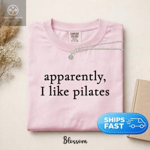 May include: A light pink crew neck sweatshirt with the text "apparently, I like pilates" in black. A silver necklace with a sun pendant is on the shirt. The brand name "Blossom" is at the bottom.