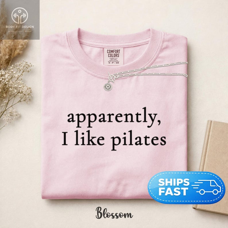 May include: A light pink crew neck sweatshirt with the text "apparently, I like pilates" in black. A silver necklace with a sun pendant is on the shirt. The brand name "Blossom" is at the bottom.