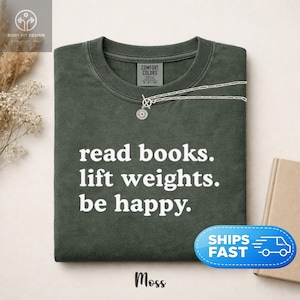 Könnte beinhalten: Ein kurzärmeliges T-Shirt in Dunkelolivgrün mit Rundhalsausschnitt. Das Shirt hat einen weißen Aufdruck auf der Vorderseite mit dem Text: read books. lift weights. be happy. Das Etikett im Nackenbereich zeigt den Markennamen Comfort Colors.
