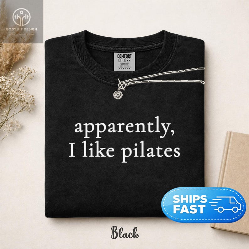 May include: Black t-shirt with the text "apparently, I like pilates" in white. A silver necklace with a sunburst pendant is draped over the shirt. The shirt has a crew neck and short sleeves.