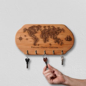 May include: Wooden key holder with a world map design. The holder has five hooks and the words "Explore, Dream, Discover" engraved below the map. A hand is reaching for a key. The holder is made of light brown wood.