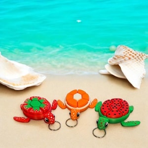 Op de afbeelding: Drie kleurrijke schildpad sleutelhangers op een zandstrand. De schildpadden zijn gemaakt van plastic en hebben een gesegmenteerd schildontwerp. De schilden zijn rood met groene accenten, oranje met witte accenten en groen met rode accenten.