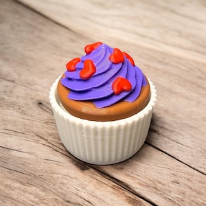 May include: A decorative cupcake with purple icing and red heart-shaped sprinkles. The cupcake sits in a white, fluted holder. The base of the cupcake is brown. The image is set against a wooden background.