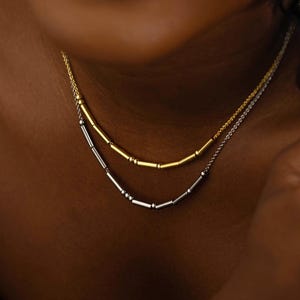 Peut inclure: Un collier superposé en or et argent avec un motif de barres. Le collier est composé de deux chaînes délicates, chacune avec une série de petites barres espacées. La chaîne supérieure est dorée et la chaîne inférieure est argentée.
