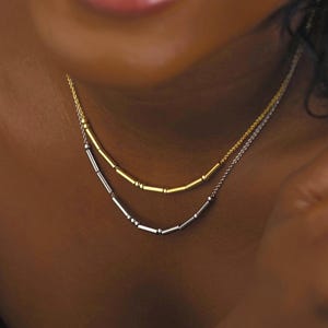 Peut inclure: Un ensemble de colliers superposés composé de deux chaînes délicates. La chaîne supérieure est dorée avec de petites perles cylindriques, tandis que la chaîne inférieure est argentée et comporte également des perles cylindriques au centre.