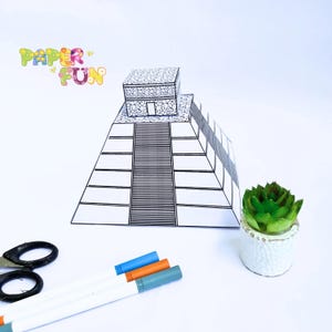 May include: A black and white paper model of a pyramid with stairs and a small building on top. The words "PAPER FUN" are in colorful bubble letters. A small green succulent in a white pot, scissors, and three markers are also present.