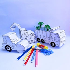 Könnte beinhalten: Ein Set aus drei Papier-Baufahrzeugen, darunter ein Kipplaster, ein Bagger und eine Walze. Die Fahrzeuge sind weiß und bereit zum Ausmalen. Das Set enthält außerdem Kleber, Schere und Stifte.