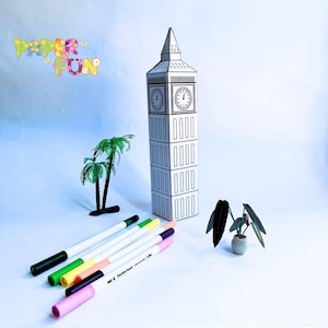 May include: A black and white paper model of Big Ben, with a clock face, is displayed next to a collection of colorful markers. The words "PAPER FUN" are written in colorful letters. A small palm tree and a potted plant are also present.