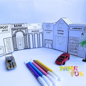 Puede incluir: Un conjunto de edificios de papel blanco con arte lineal negro que incluye oficina de correos, banco, museo, clínica veterinaria, clínica dental y biblioteca. Dos coches de juguete y cuatro rotuladores de colores están dispuestos delante.