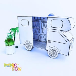 Op de afbeelding: Een wit papieren model van een camper met de deur open, waardoor een eenvoudig interieur met meubels zichtbaar wordt. Het model staat op een wit oppervlak met een kleine plant in een pot op de voorgrond.