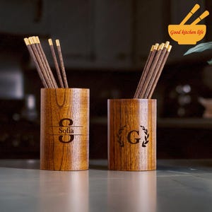 Peut inclure: Deux porte-baguettes en bois avec des gravures personnalisées. Un porte-baguettes porte le nom "Sofia" gravé, et l'autre porte la lettre "G" avec un motif de laurier. Les deux porte-baguettes sont en bois foncé avec un motif de grain naturel.