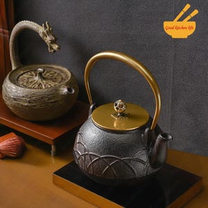 Peut inclure: Deux théières en fonte avec des accents dorés. La théière de gauche a un bec en forme de dragon et une finition brune. La théière de droite a une finition noire avec un motif décoratif et un couvercle doré. Le texte "Good kitchen life" est visible dans le coin supérieur droit de l'image.