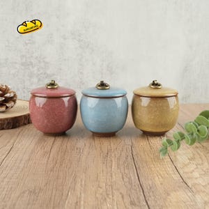 May include: Three ceramic jars with lids in red, blue, and yellow. Each jar has a crackle glaze finish and a small, decorative knob on the lid. The jars are arranged on a wooden surface.
