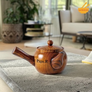 May include: A wooden teapot with a dark brown finish, featuring a spout, handle, and a lid with a small knob. The text "Papa.R" is printed on the side. The teapot is sitting on a gray patterned surface. The words "Good kitchen life" are in the top right corner.