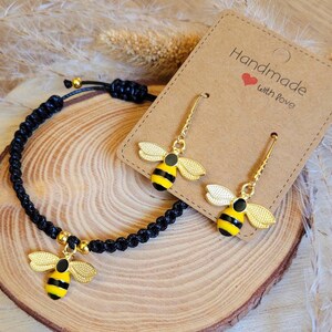 Könnte beinhalten: Ein schwarzes geflochtenes Armband und goldfarbene Bienenohrringe mit gelben und schwarzen Streifen werden auf einer Holzoberfläche präsentiert. Die Ohrringe und das Armband haben einen Bienenanhänger. Eine braune Karte mit dem Text "Handmade with Love" ist ebenfalls vorhanden.