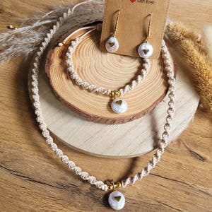 Op de afbeelding: Een handgemaakte sieradenset met een ketting, armband en oorbellen. De set is gemaakt van beige macramé koord met goudkleurige accenten en parelmoer hangers met gouden hartdetails. De sieraden worden gepresenteerd op een houten oppervlak.
