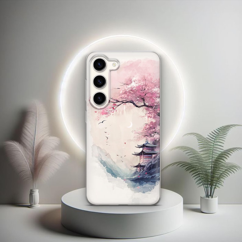 Puede incluir: Una funda transparente para tel&eacute;fono con un dise&ntilde;o de acuarela de un cerezo rosa en flor y un pagodo en el fondo. La funda tiene un recorte para una c&aacute;mara de triple lente.