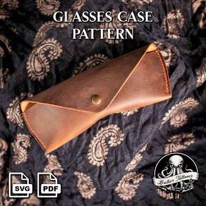 May include: A brown leather glasses case with a button closure. The case is displayed on a dark fabric with a paisley pattern. The text "GLASSES CASE PATTERN" is at the top. SVG and PDF icons are in the lower left corner.