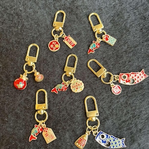 May include: Six gold keychains with different charms. The charms include a red and gold fish, a red and gold pomegranate, a gold coin with Chinese characters, a gold and red tag with Chinese characters, a gold and blue carp, and a gold and blue tag with Chinese characters. The keychains are on a grey background.