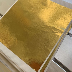 Puede incluir: Una pila de hojas de pan de oro, cada hoja está protegida por un respaldo de papel blanco. La hoja de oro es de un color dorado brillante y brillante.