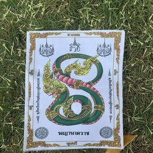 Könnte beinhalten: Ein weißer Stoff mit goldener Umrandung und einem grünen und goldenen Schlangendesign. Die Schlange ist zu einem Kreis zusammengerollt und hat eine rote Zunge. Der Stoff trägt den Text "พญานาคราช" in thailändischer Schrift.