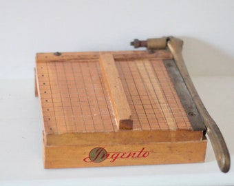May include: Vintage wooden paper cutter with a grid pattern and a metal blade. The paper cutter has a wooden base with the word "Ingento" in red script. The blade is attached to a metal arm.