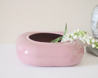 May include: A pink ceramic vase with a rounded, oblong shape. The interior is dark brown. White flowers with green stems are arranged in the vase. A silver bust is in the background.