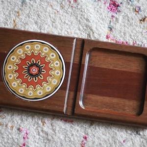 May include: Wooden serving tray with a decorative ceramic coaster. The coaster features a floral design in orange, yellow, and black. The tray has a rectangular cutout and a slot for holding items. The wood has a warm, reddish-brown tone.