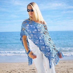 Pode incluir: Um xale de crochê azul claro com um padrão floral e geométrico. O xale tem franjas na borda inferior e é usado sobre uma blusa branca e calças brancas. O fundo é uma praia com o oceano e o céu azul.