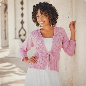 May include: A light pink knitted cardigan with a textured pattern on the front and sleeves. The cardigan has a tie closure and scalloped edges. The sleeves are three-quarter length, and the garment is worn over a white dress.