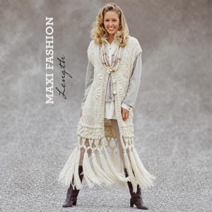 May include: A cream-colored sleeveless long cardigan featuring an intricate cable knit pattern, a button-down front, and a long fringe hem. The garment is worn over a white shirt and light-colored pants, paired with brown boots and a beaded necklace.