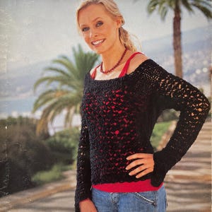 May include: A black crochet sweater with long sleeves and an open weave design. The sweater is worn over a red top and blue jeans. A red beaded necklace is visible. The background features palm trees and a coastal view.