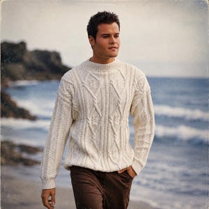 May include: Cream-colored cable-knit sweater with a crew neck. The sweater features intricate woven patterns and is worn with brown pants. The person is walking on a beach with the ocean in the background.