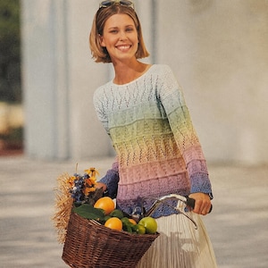 May include: A rainbow-colored knit sweater with a crew neck and long sleeves. The sweater transitions from light blue to pink, with yellow and green hues in between. The woman is holding a bicycle with a basket filled with fruit and flowers.