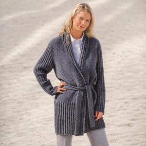 May include: A charcoal gray knit cardigan featuring a shawl collar, long sleeves with ribbed detailing, and a matching tie belt at the waist. The garment is worn over a crisp white collared shirt and light gray trousers, creating a layered look.