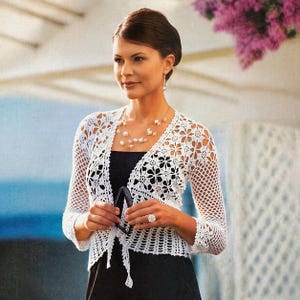 May include: A white, open-front crochet cardigan with a floral pattern. The cardigan has long sleeves and a tie closure. The woman is wearing a black dress and a pearl necklace. She is also holding a pair of sunglasses.